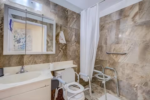 Bathroom with beige marble-patterned walls, a white sink with a mirror cabinet above it, a white shower curtain, and a white shower chair with armrests inside the shower area. There is also a grab bar mounted on the shower wall.