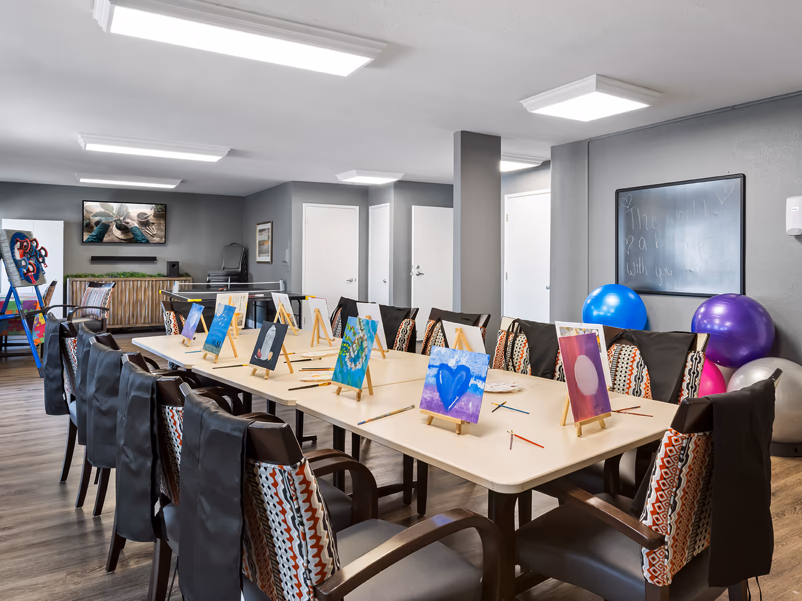 A well-lit activity room with a long table surrounded by chairs, each with a small easel holding a colorful painting. Paintbrushes and palettes are scattered on the table. In the background, there is a TV mounted on the wall, exercise balls, a blackboard with writing, and some stacked chairs.
