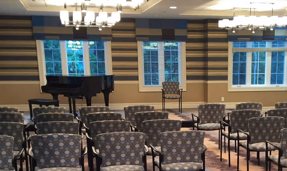 A room with rows of patterned chairs facing a small stage area with a single chair and a black grand piano. The walls have horizontal striped wallpaper and three large windows with white frames, allowing natural light to enter.