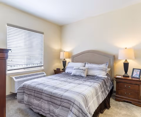 A cozy bedroom with a neatly made bed featuring a gray and white striped comforter and a decorative pillow that says 'welcome HOME'. The bed has a cushioned headboard and is flanked by two wooden nightstands, each with a lamp. A window with blinds and an air conditioning unit is on the left side of the room.