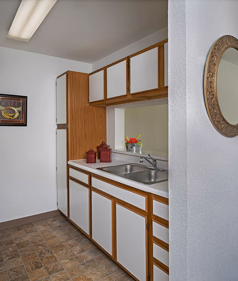 A small kitchen area with white and wood-trimmed cabinets, a double stainless steel sink, two red canisters on the counter, a small flower arrangement in a metal pot, a framed picture of a coffee cup on the wall, and a decorative round mirror on an adjacent wall.