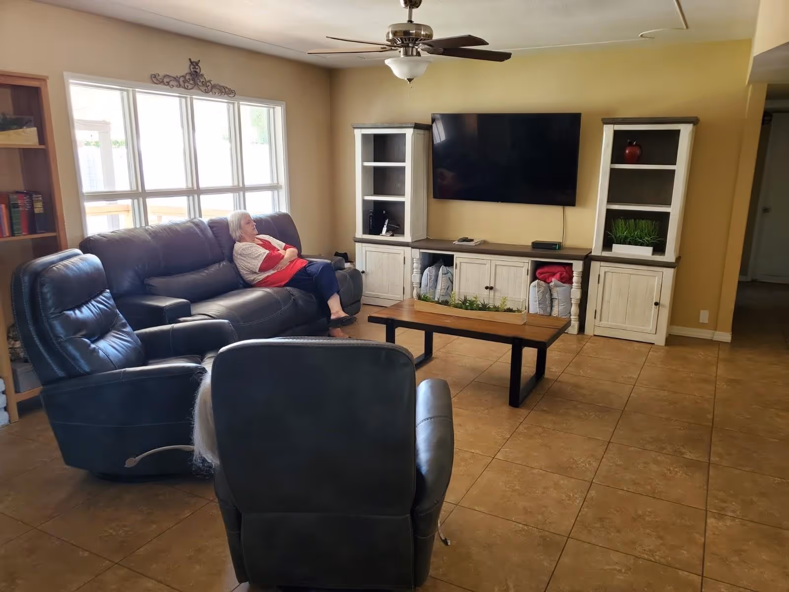 A living room with a large window letting in natural light. There is a woman sitting on a dark leather couch, with two matching recliners in the foreground. A wooden coffee table with a planter sits in the center. Against the wall is a mounted flat-screen TV flanked by two white wooden cabinets with shelves holding decorative items. The floor is tiled and the walls are painted a warm yellow color.