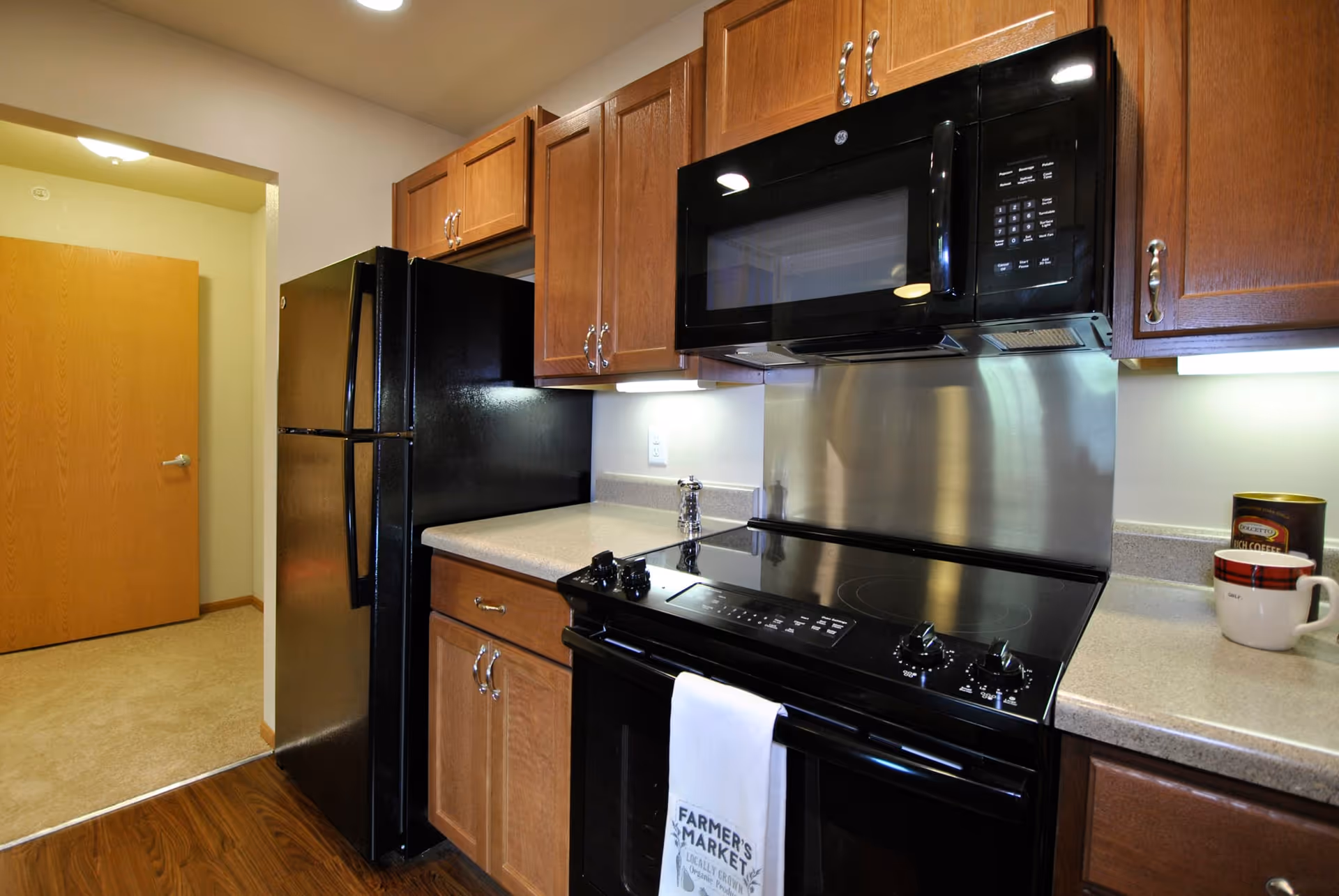 A kitchen area featuring wooden cabinets, a black refrigerator, a black microwave mounted above a black electric stove with a white towel hanging on the oven handle, and a countertop with a coffee mug and a container. The floor transitions from wood in the kitchen to carpet in the adjacent room with a wooden door.