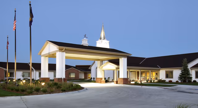 Exterior view of Brookefield Park Skilled Nursing & Rehabilitation facility at dusk, showing a covered entrance with white columns and brick bases, three flagpoles with flags, well-maintained landscaping, and a building with a steeple and lit windows.