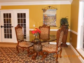 A dining area with a round glass table and four ornate wooden chairs with patterned upholstery. The room has yellow walls with white trim, a decorative painting hanging on the wall, a potted plant on a wooden pedestal, and double French doors with glass panes. The floor is covered with a patterned carpet.