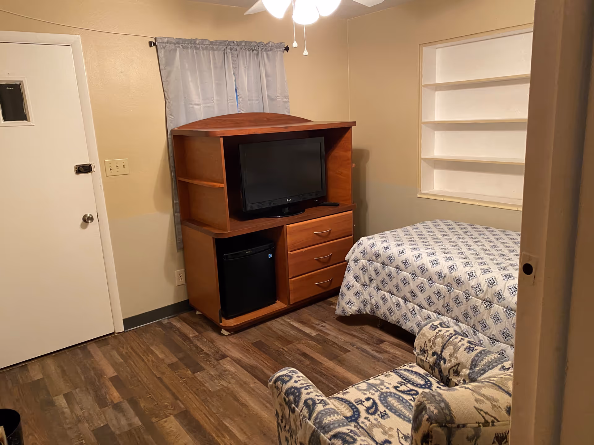 A small bedroom with a single bed covered in a patterned blue and white comforter, a wooden TV stand with a flat-screen TV and a mini refrigerator, a patterned armchair, a window with gray curtains, and built-in white shelves on the wall.