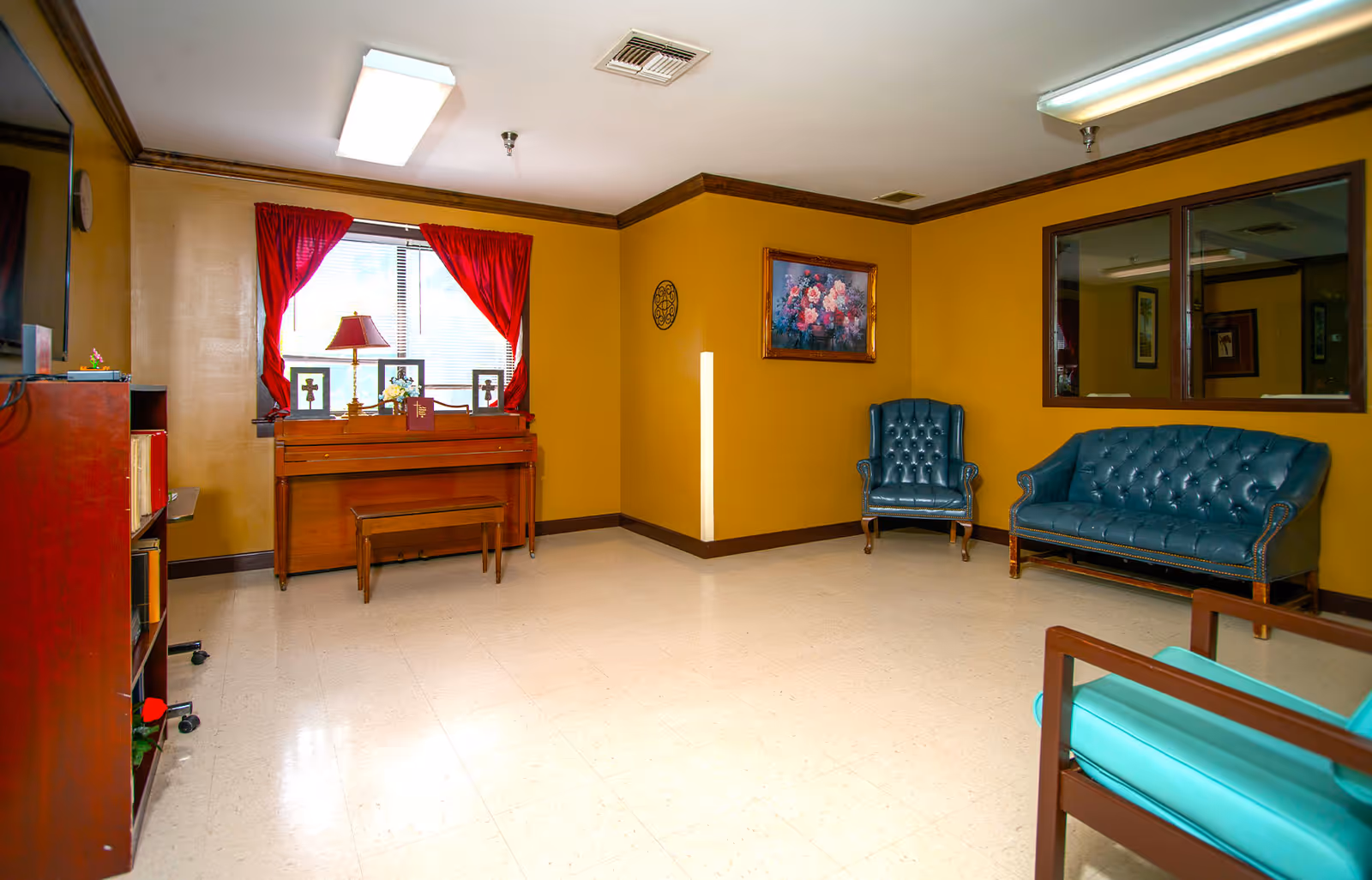 A cozy room with mustard yellow walls and a white tiled floor featuring a wooden piano with a bench in front of a window with red curtains. There is a table lamp and framed pictures on the piano. The room also has a blue leather armchair, a matching blue leather sofa, a wooden bookshelf, and a framed floral painting on the wall. A large window with a wooden frame looks into another room.