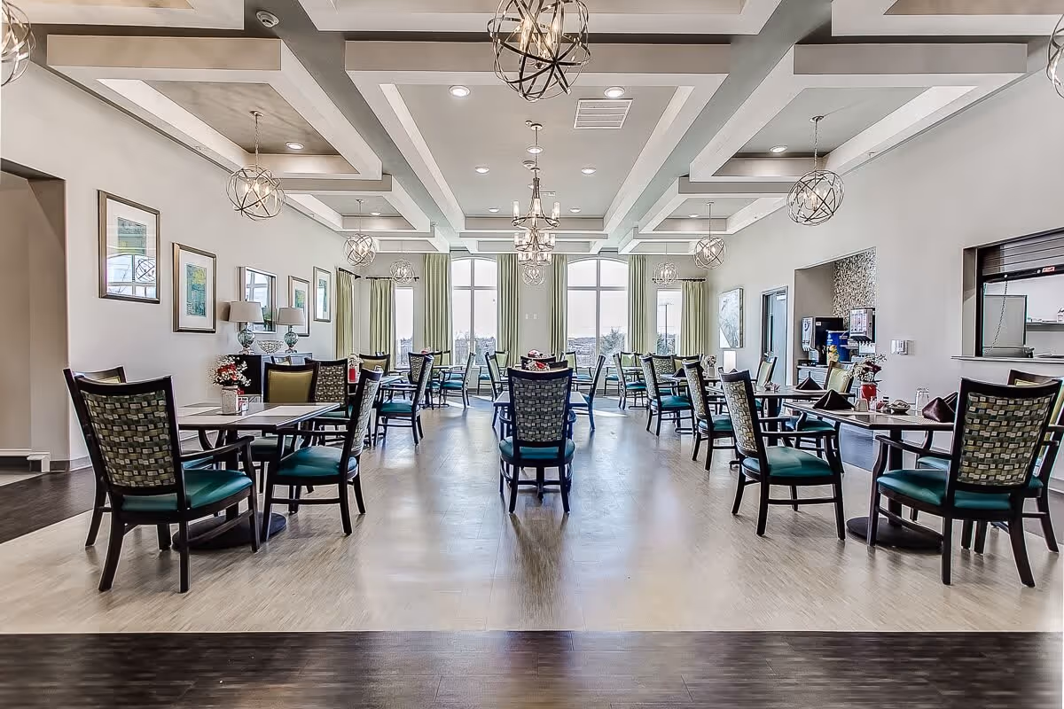 A spacious and well-lit dining room in a senior living facility with multiple tables and chairs arranged neatly. The room features large windows with green curtains, modern chandeliers hanging from a coffered ceiling, framed artwork on the walls, and a serving area with a coffee machine on the right side.
