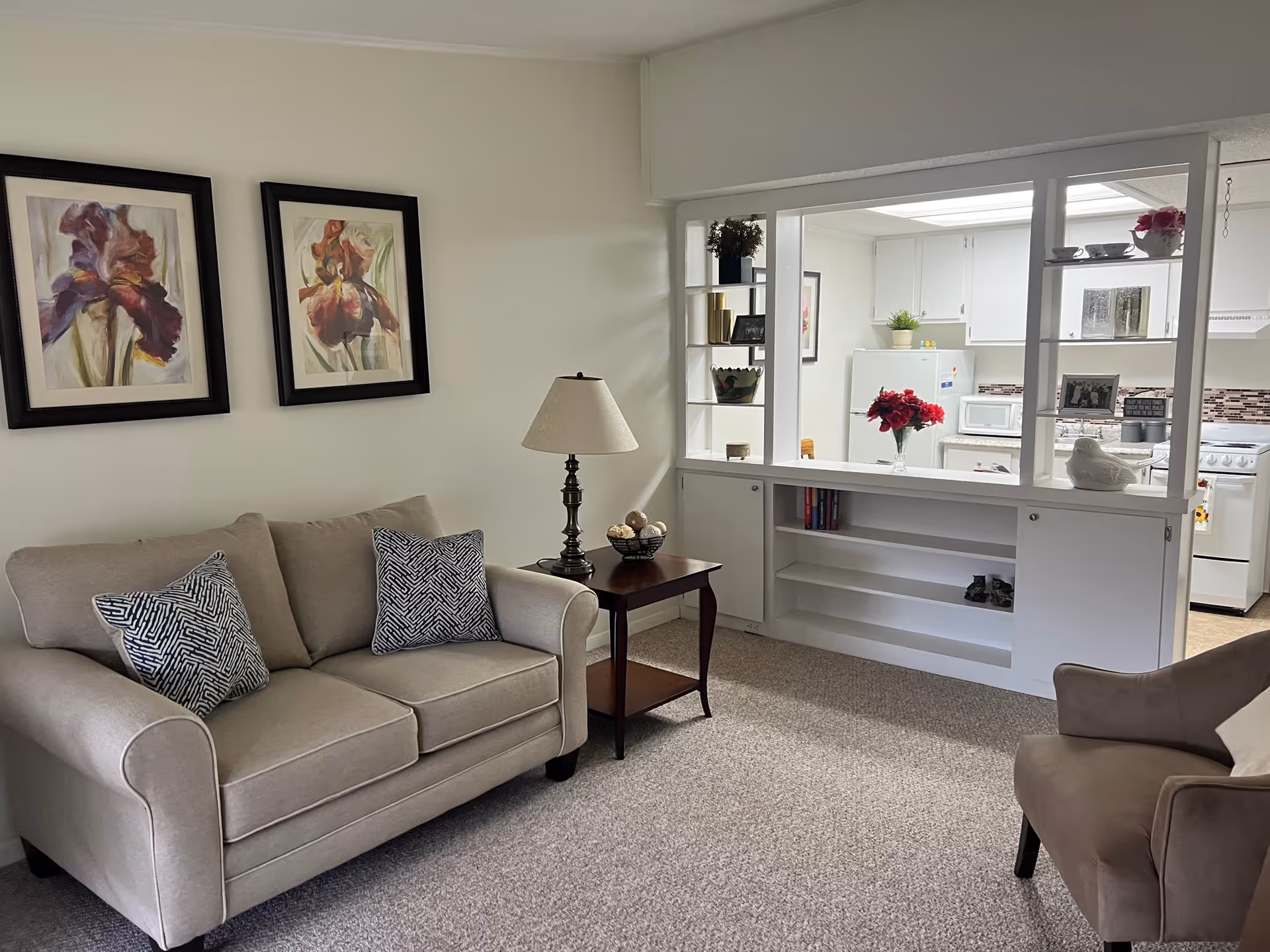 A cozy living room with a beige loveseat adorned with two patterned pillows, a wooden side table with a lamp and decorative bowl, and a single armchair. The room features two framed floral paintings on the wall and a large open window-like partition that looks into a white kitchen with cabinets, a refrigerator, microwave, and stove. The kitchen counter has a vase with red flowers and some decorative items on shelves.