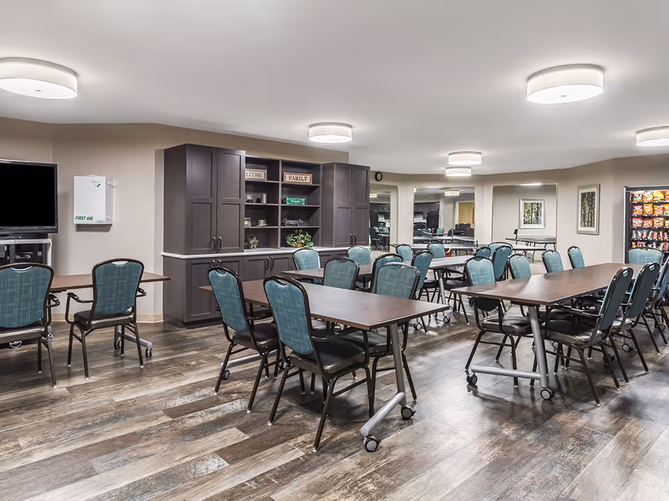 A spacious room with multiple rectangular tables and teal cushioned chairs arranged around them, a large cabinet with shelves and decorative items, a vending machine, a wall-mounted TV, and a first aid box. The room has wood-patterned flooring and bright ceiling lights.