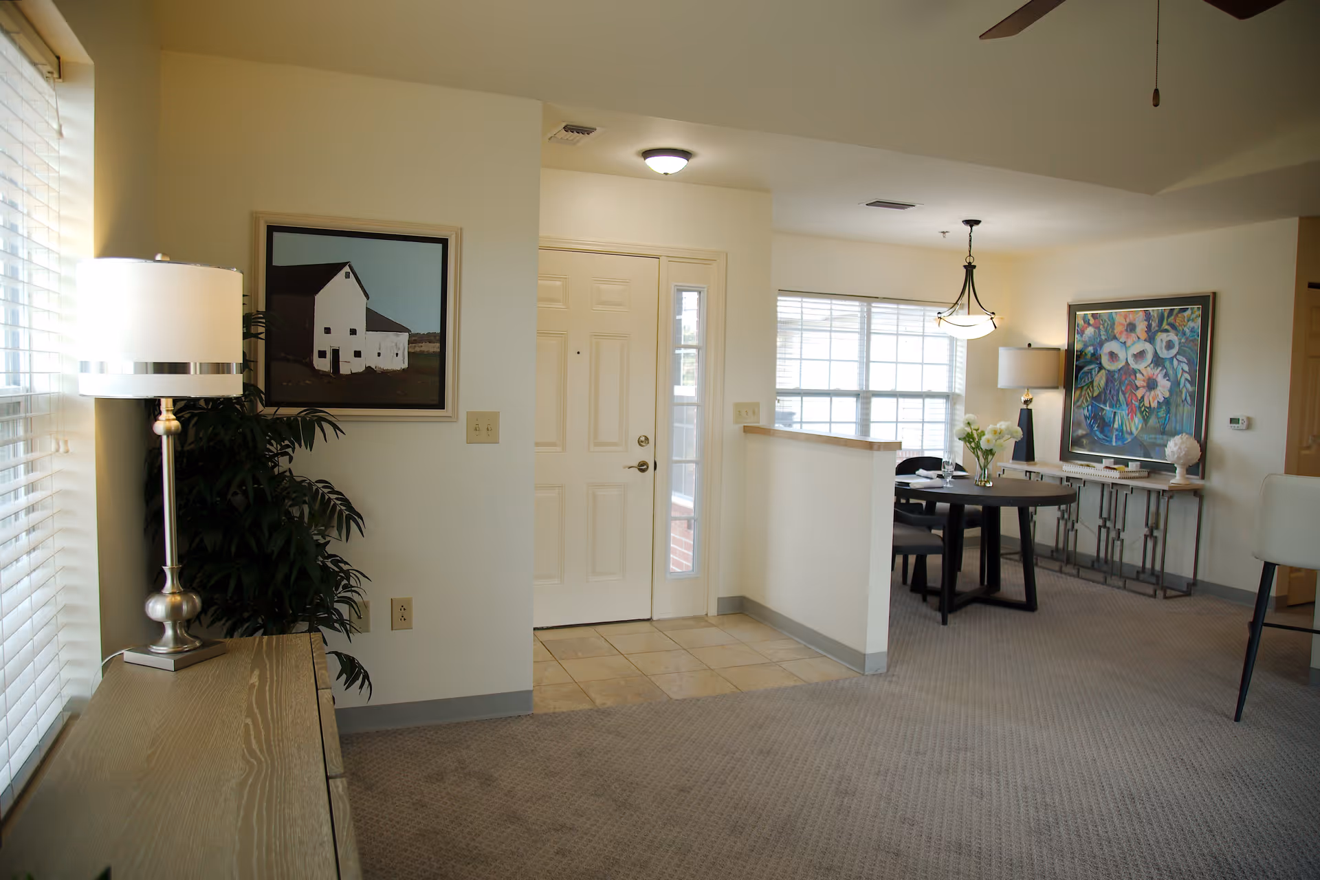 Interior view of a senior living facility showing an entryway with a white door and a small tiled floor area. To the left, there is a wooden console table with a silver lamp and a potted plant. A framed picture of a barn hangs on the wall above the plant. To the right, there is a dining area with a round black table, four chairs, a hanging light fixture, and a console table against the wall with a colorful floral painting above it. The room is carpeted and well-lit with natural light from large windows.