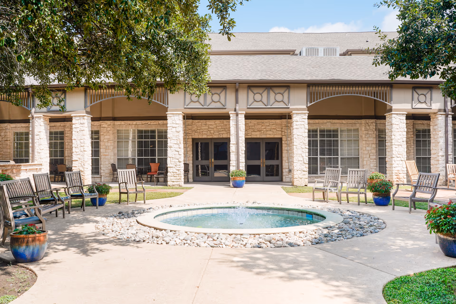Outdoor courtyard area at The Reserve at North Dallas Assisted & Senior Living featuring a circular water fountain surrounded by rocks, multiple chairs arranged around the fountain, potted plants, and a building with stone pillars and large windows in the background under a clear blue sky.