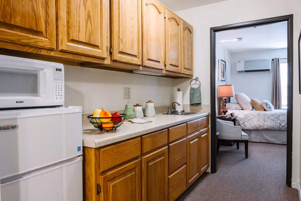 A small kitchenette with wooden cabinets, a white microwave on top of a mini refrigerator, a countertop with a fruit basket and kitchen containers, and a sink. Through an open doorway, a bedroom is visible with a bed, pillows, a bedside table with a lamp, a chair, and an air conditioning unit on the wall.
