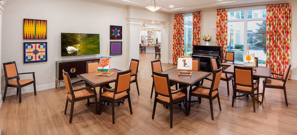 A bright and spacious common area with wooden floors, several tables and chairs arranged for group activities. Each table has an easel with a painting on it. The walls are decorated with colorful geometric artwork and a flat-screen TV displaying an image of a swan on a pond. Large windows with red and yellow patterned curtains let in natural light and offer a view of the outdoor courtyard. A black piano is positioned near the windows.