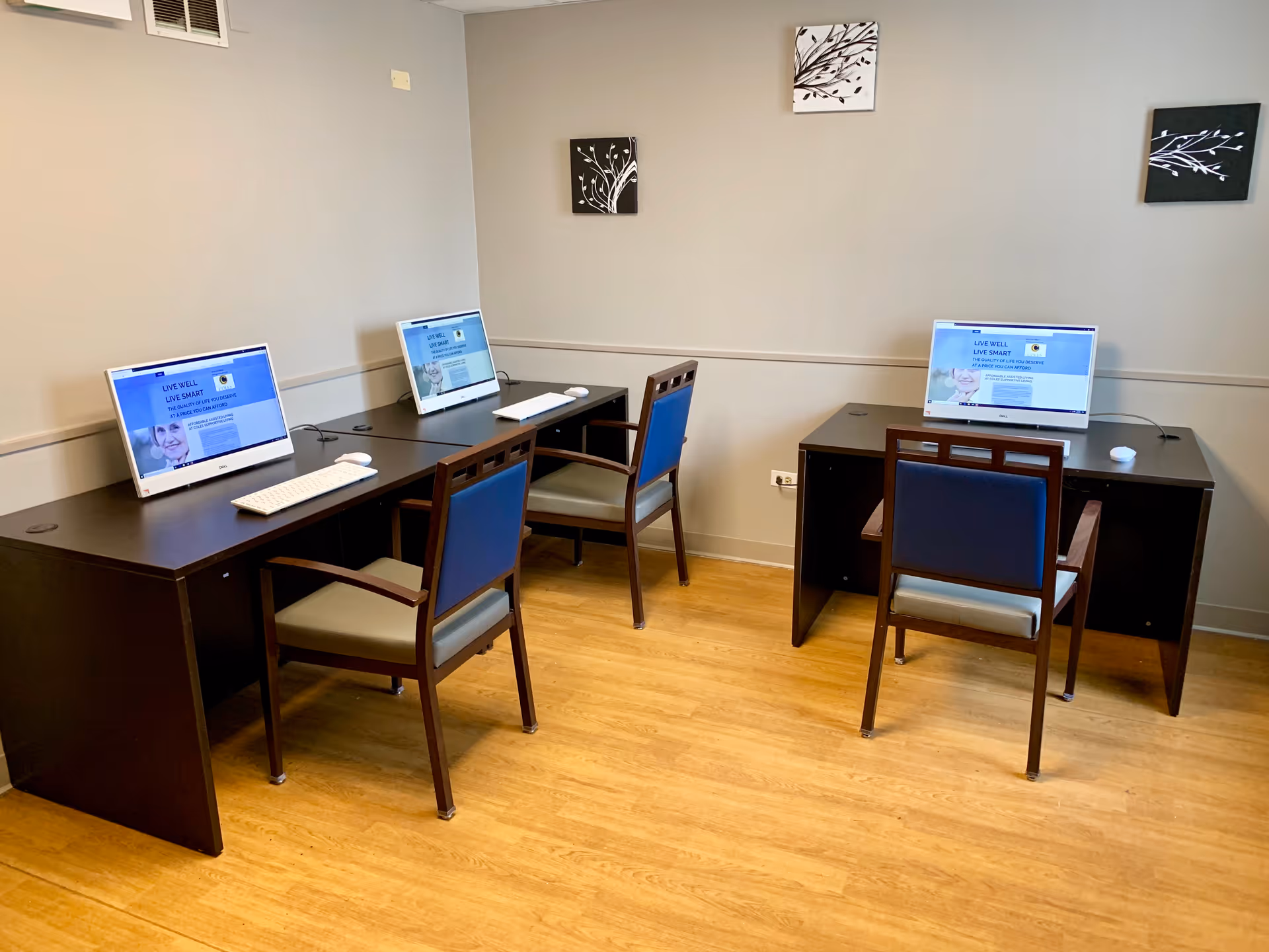 A small computer room with three desks, each equipped with a desktop computer, keyboard, and mouse. Three chairs with blue backs and armrests are positioned at the desks. The room has light-colored walls with three decorative black and white wall hangings and a wooden floor.