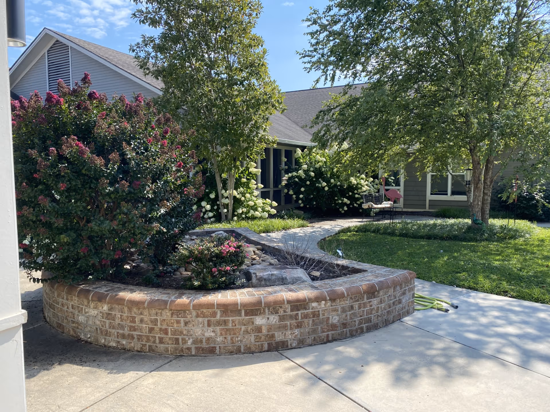 Outdoor garden area at Concord Place Memory Care featuring a curved brick planter with various flowering bushes and small trees, a concrete walkway, green grass, and a building with windows in the background under a clear blue sky.