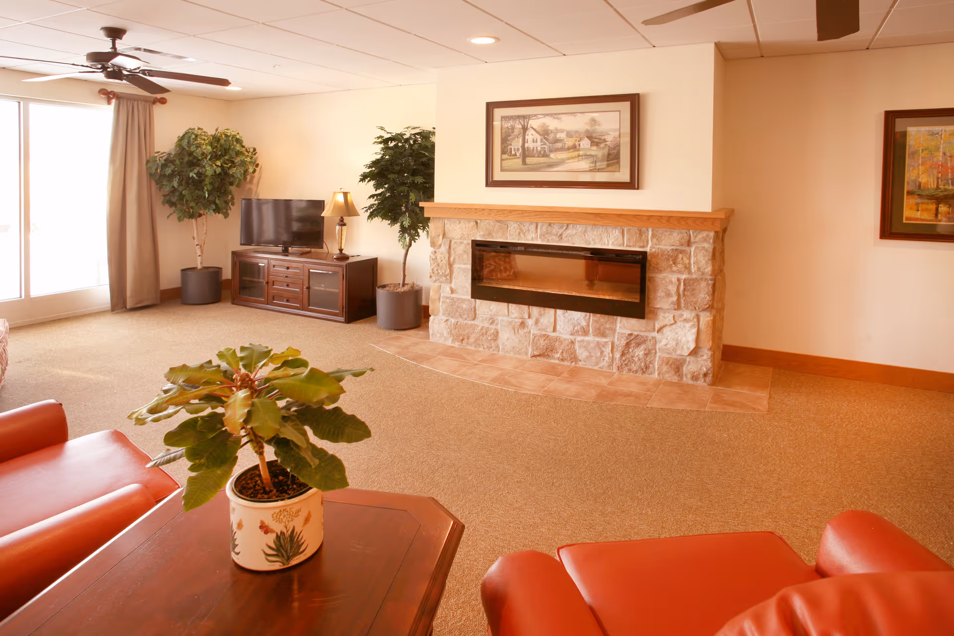 A cozy living room with a stone fireplace, a flat-screen TV on a wooden stand, two potted plants, framed artwork on the walls, a ceiling fan, and red leather chairs around a wooden coffee table with a potted plant on it.