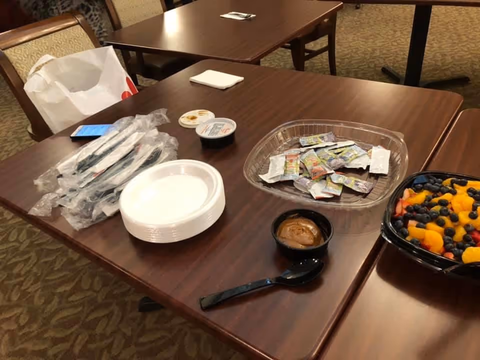 Dining tables with disposable plates, wrapped utensils, condiment packets and a container of mixed fruit in a communal dining area.