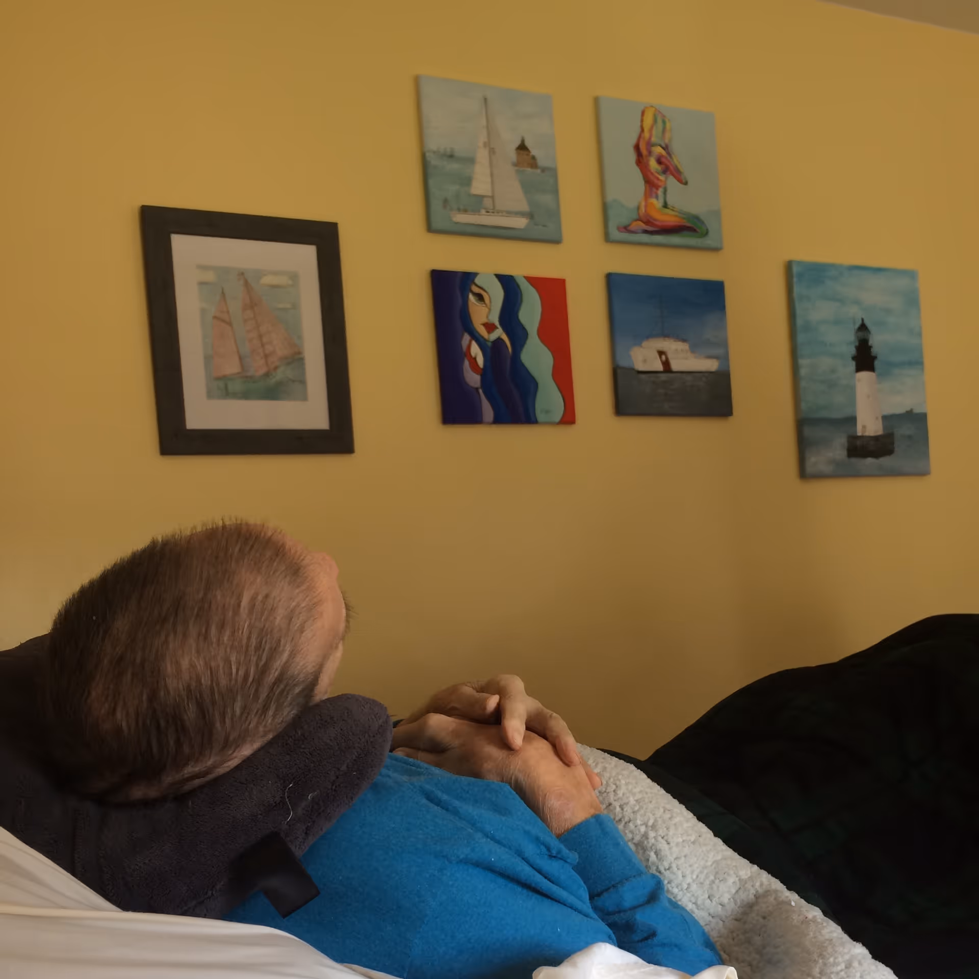 A person lying in bed facing a yellow wall decorated with several framed nautical-themed paintings.