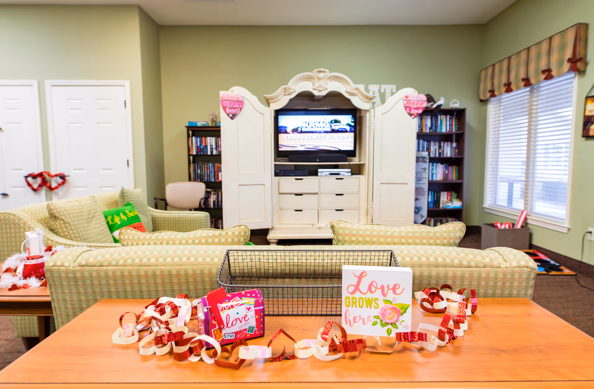 A cozy living room area in an assisted living facility with green patterned sofas facing a white entertainment center holding a TV and drawers. The room is decorated with Valentine-themed paper chains and signs, including one that says 'Love grows here.' Bookshelves flank the entertainment center, and a window with blinds and a valance is on the right wall.