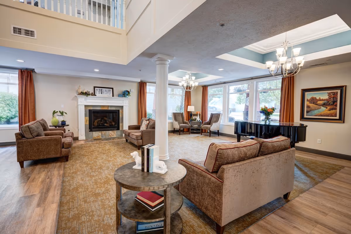 A spacious and well-lit living room area in South Hill Village featuring comfortable brown sofas and armchairs arranged around a fireplace. The room has large windows with orange curtains, a grand piano, a round table with books, and two chandeliers hanging from the ceiling. There is a painting on the wall and a column in the center of the room.