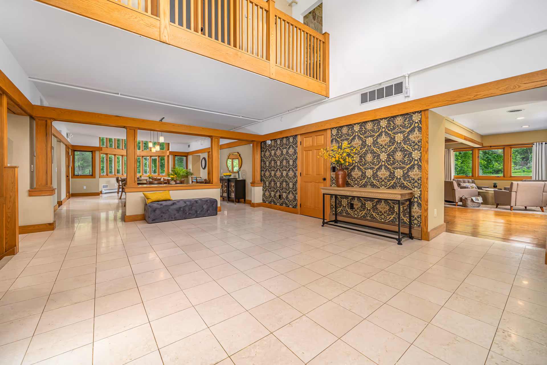 A spacious lobby area featuring a stylish design with a high ceiling, wooden accents, and large windows. A gray bench sits in the foreground, while a decorative table with a vase of flowers is positioned against a patterned wall. The room is well-lit and opens to a dining area in the background.