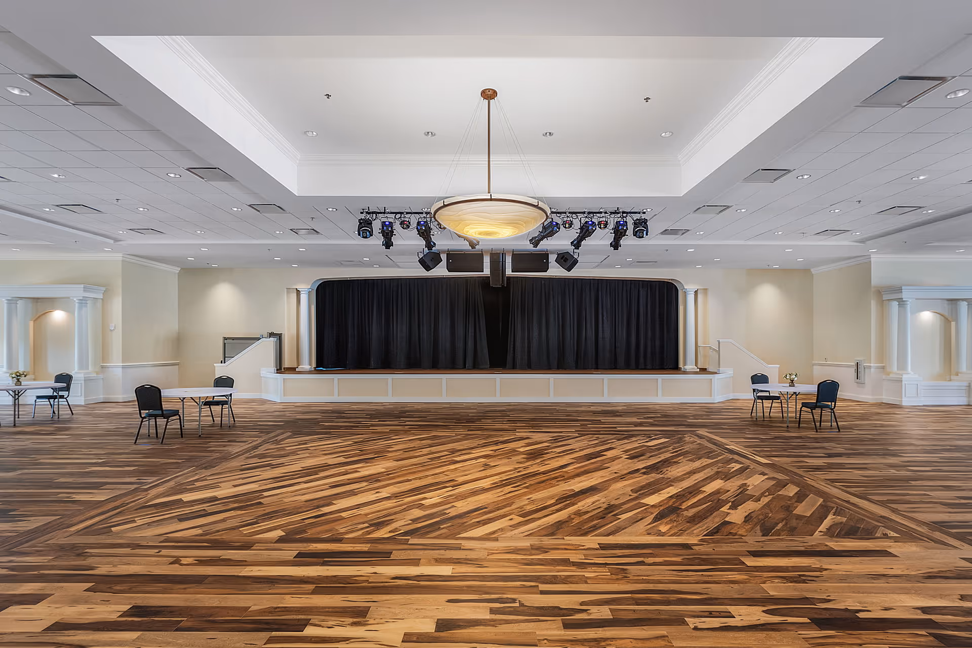 A large, empty event room with polished wooden floors and a small stage with black curtains at the far end. The ceiling has recessed lighting and a central chandelier. There are a few tables and chairs spaced around the room, and the walls are painted light beige with white trim and decorative columns.