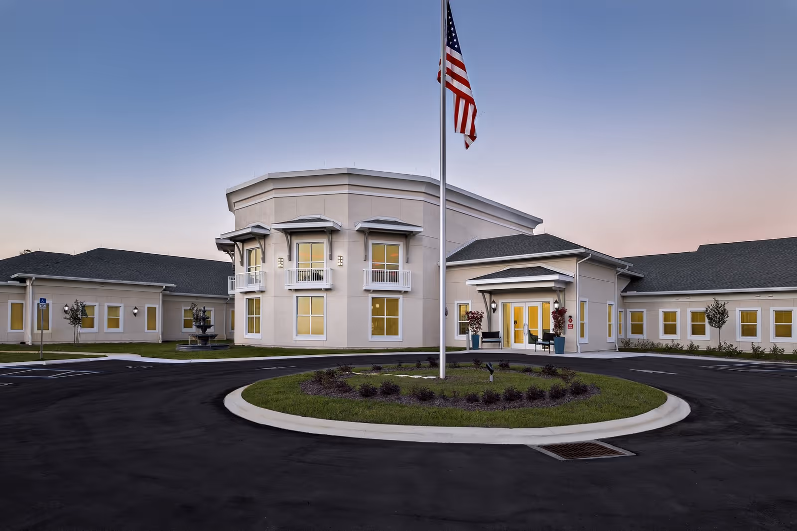 Exterior front view of Magnolia Trails at East Lake facility during dusk, showing a circular driveway with a flagpole and American flag in the center, beige building with multiple windows, small balconies, and entrance doors with potted plants on either side.
