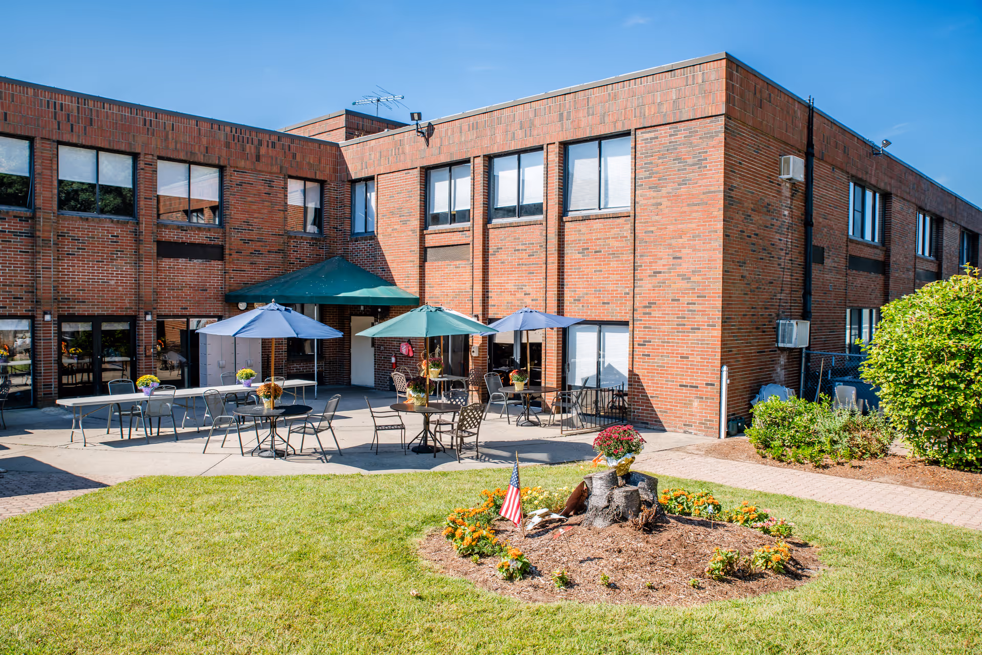 Outdoor patio area of a brick senior living facility building with several tables and chairs under umbrellas, surrounded by a well-maintained lawn and flower bed with an American flag.