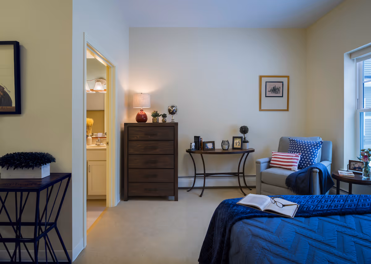 A tidy bedroom with a bed in the foreground, a dresser with a lamp, a side table and armchair by a window, and an open doorway to a bathroom.