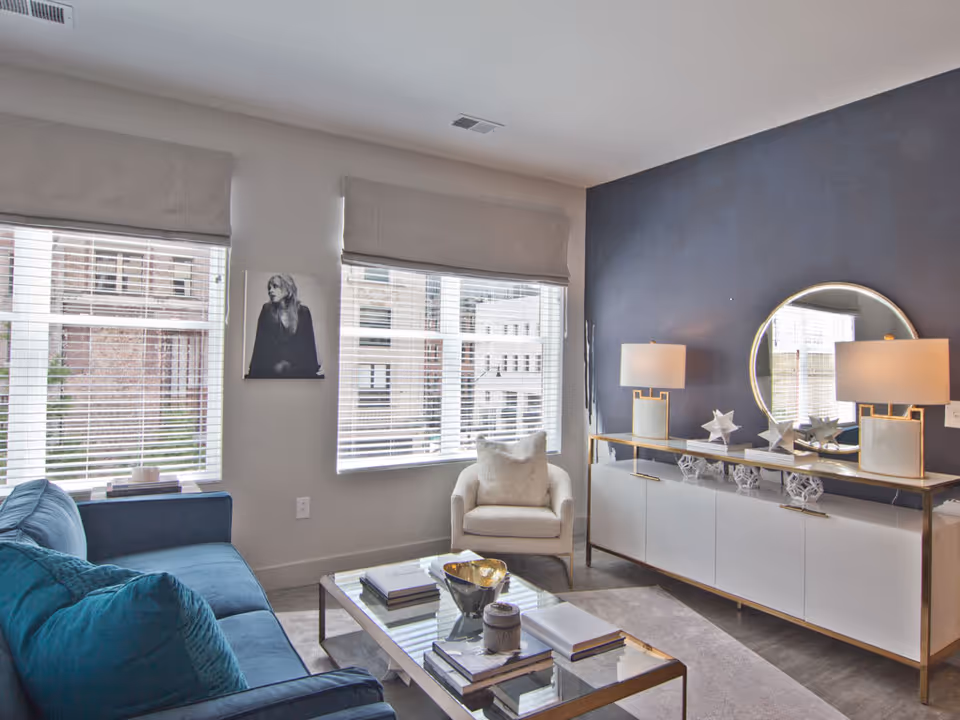 Bright modern living room with a blue sofa, armchair, glass coffee table, and a sideboard topped with lamps and a round mirror.