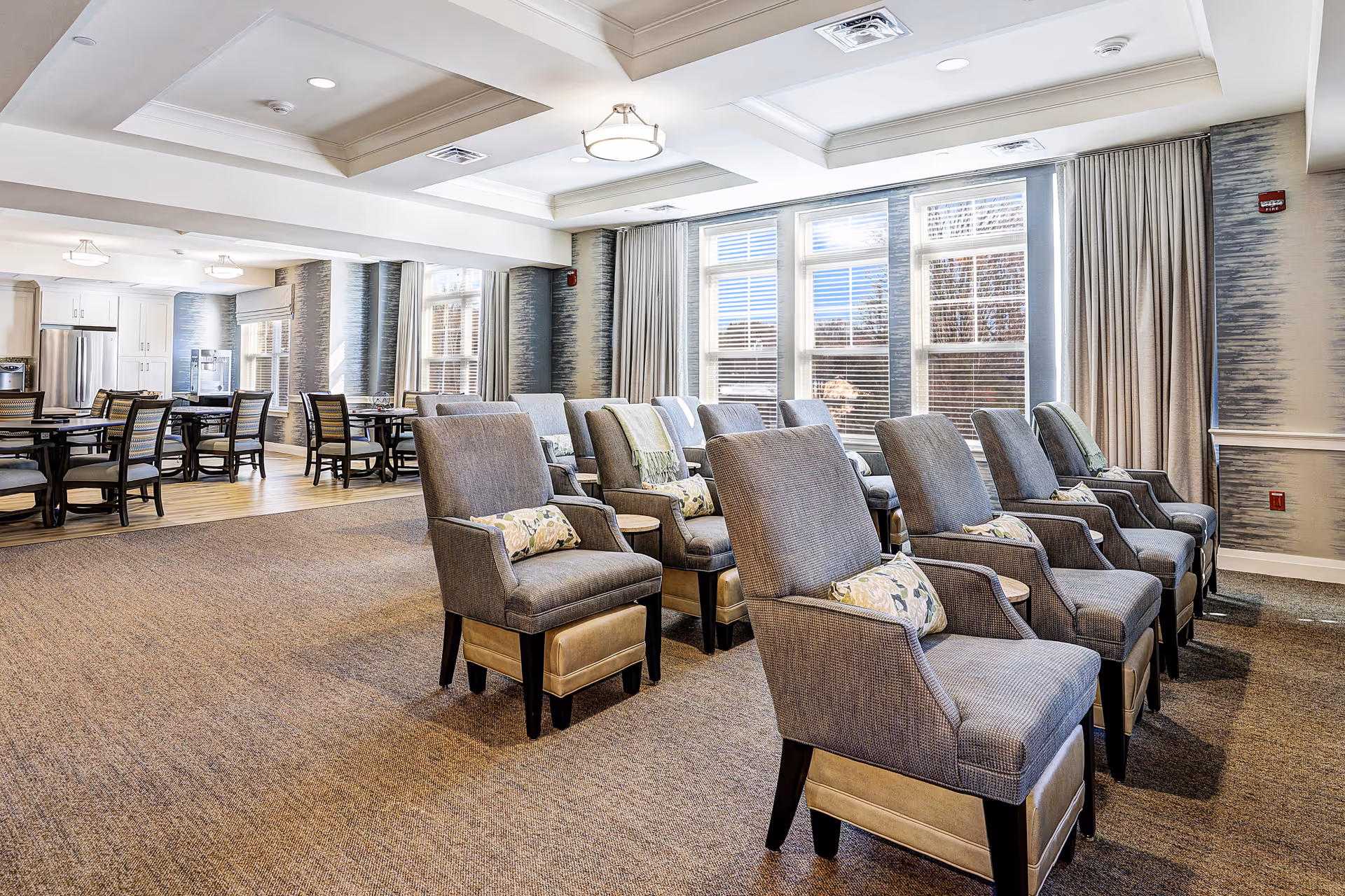 A spacious and well-lit common area in a senior living facility featuring multiple rows of comfortable upholstered chairs with decorative pillows, large windows with curtains allowing natural light to fill the room, and a dining area with tables and chairs in the background near a kitchen area with stainless steel appliances.