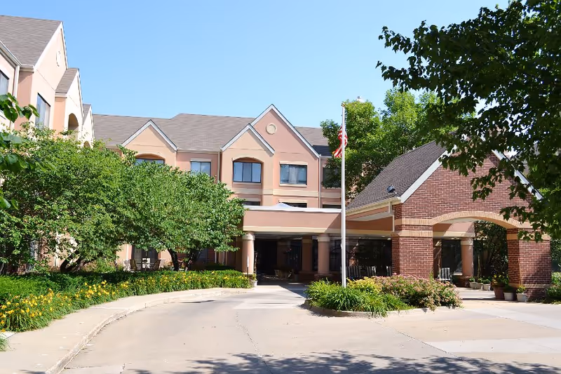 Front entrance of a multi-story retirement campus building with a covered driveway, landscaping, and an American flag.