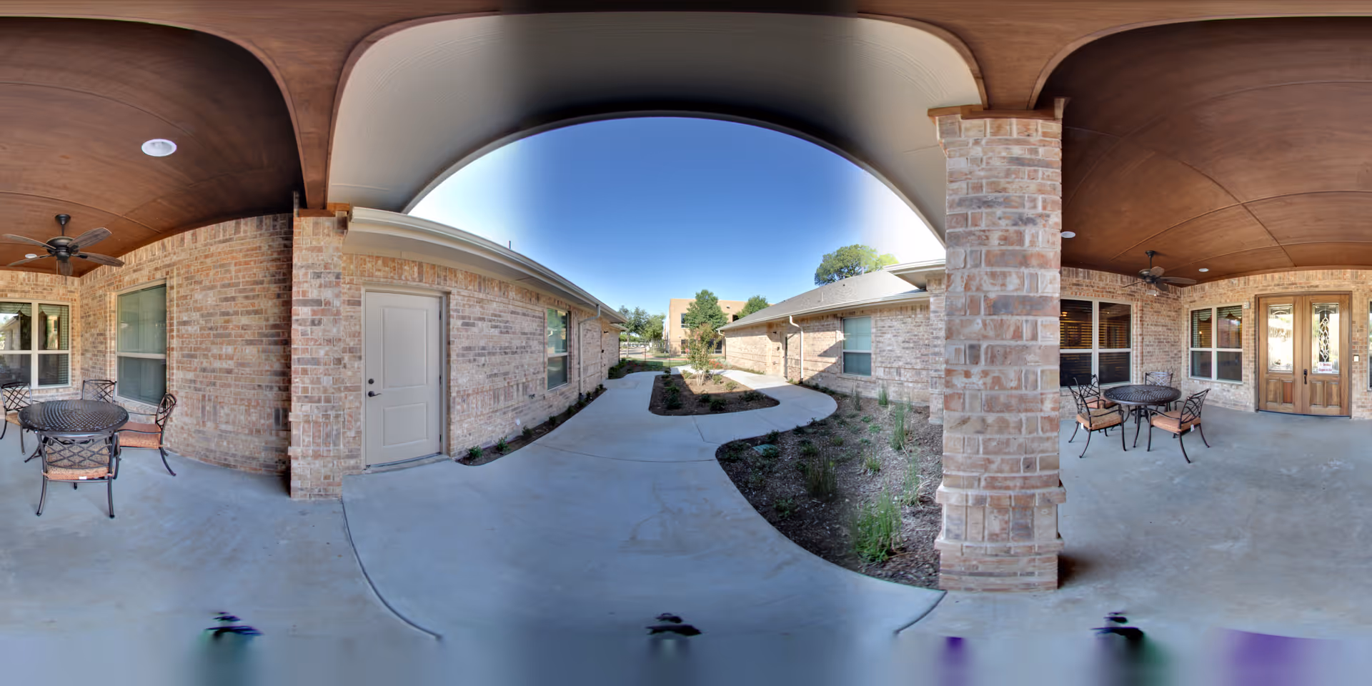 Covered outdoor patio area with brick columns and walls, featuring metal tables and chairs with cushions. The patio overlooks a concrete walkway and landscaped garden beds with small plants. The sky is clear and blue.