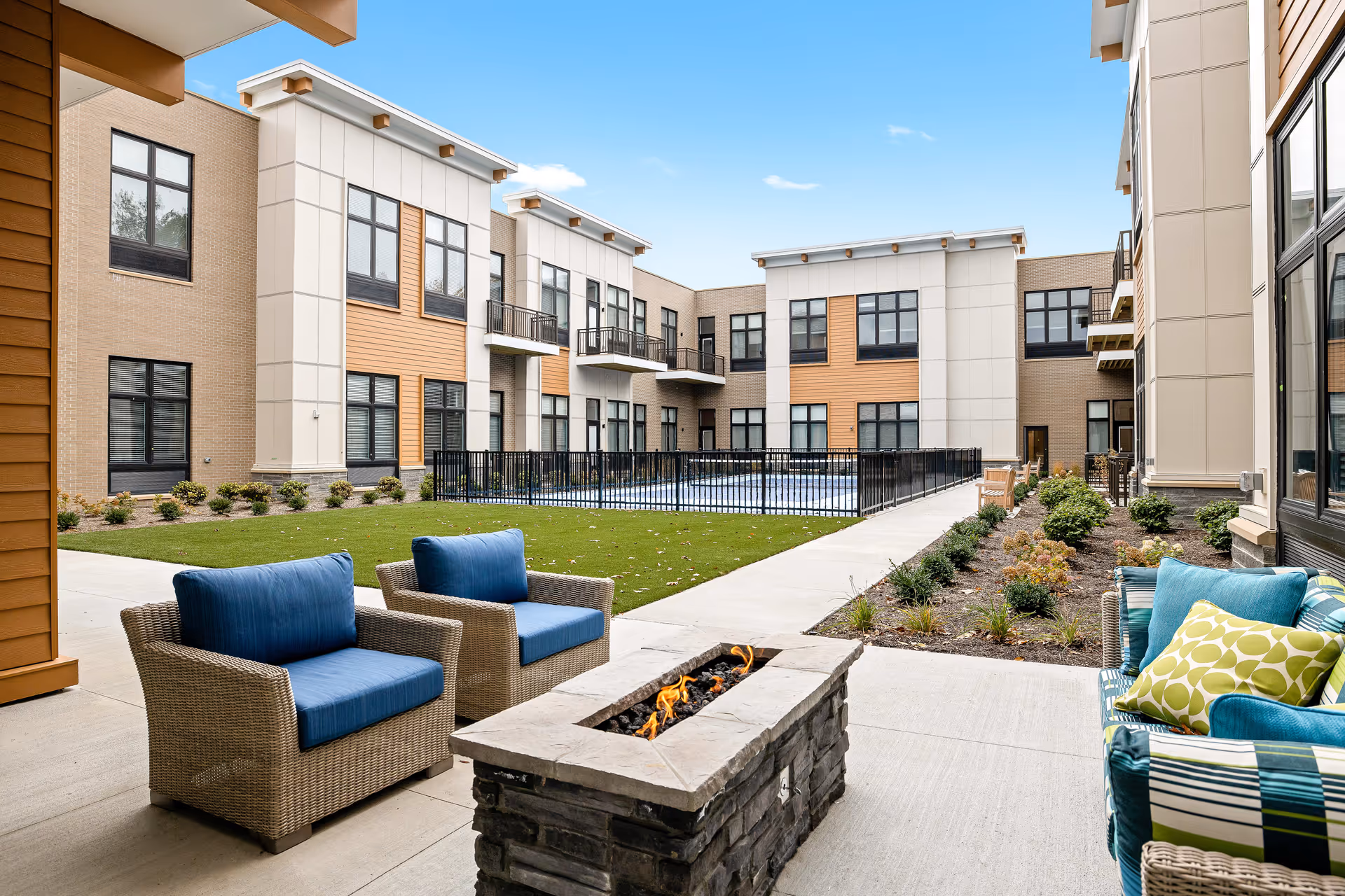 Outdoor patio area at The Ashton at Mayfield Heights featuring wicker chairs with blue cushions, a stone fire pit with flames, a cushioned wicker sofa with patterned pillows, a grassy lawn, and a modern two-story building with large windows and balconies under a clear blue sky.