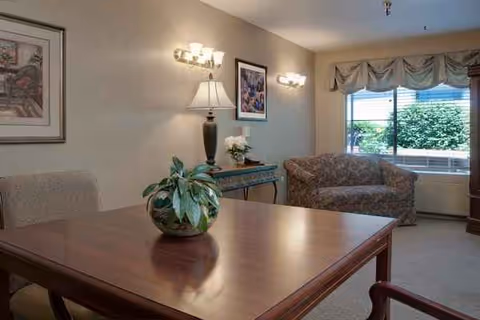 A senior living common area featuring a wooden dining table, plants, and seating by a window.