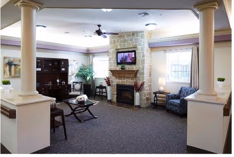 Spacious common living room with a stone fireplace and mounted TV, seating areas, coffee table, bookshelf, and decorative columns.