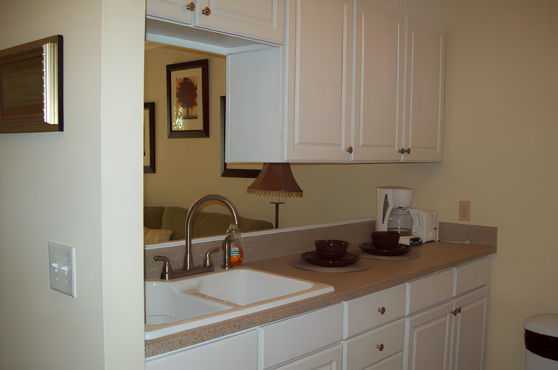 A kitchen area with white cabinets, a double sink with a faucet, a coffee maker, and two place settings with brown bowls and plates on the countertop. A mirror above the sink reflects part of a living room with a lamp and framed pictures on the wall.