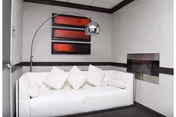 A modern living room area featuring a white tufted sofa with five white cushions, a curved floor lamp with a metallic shade, three rectangular red and black abstract wall art pieces, and a built-in wall fireplace.