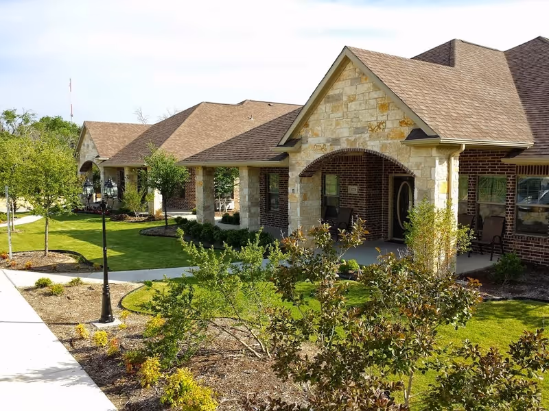 Exterior view of a single-story assisted living facility with a stone and brick facade, covered porch areas with chairs, landscaped garden beds, small trees, and a paved walkway with a vintage-style street lamp.