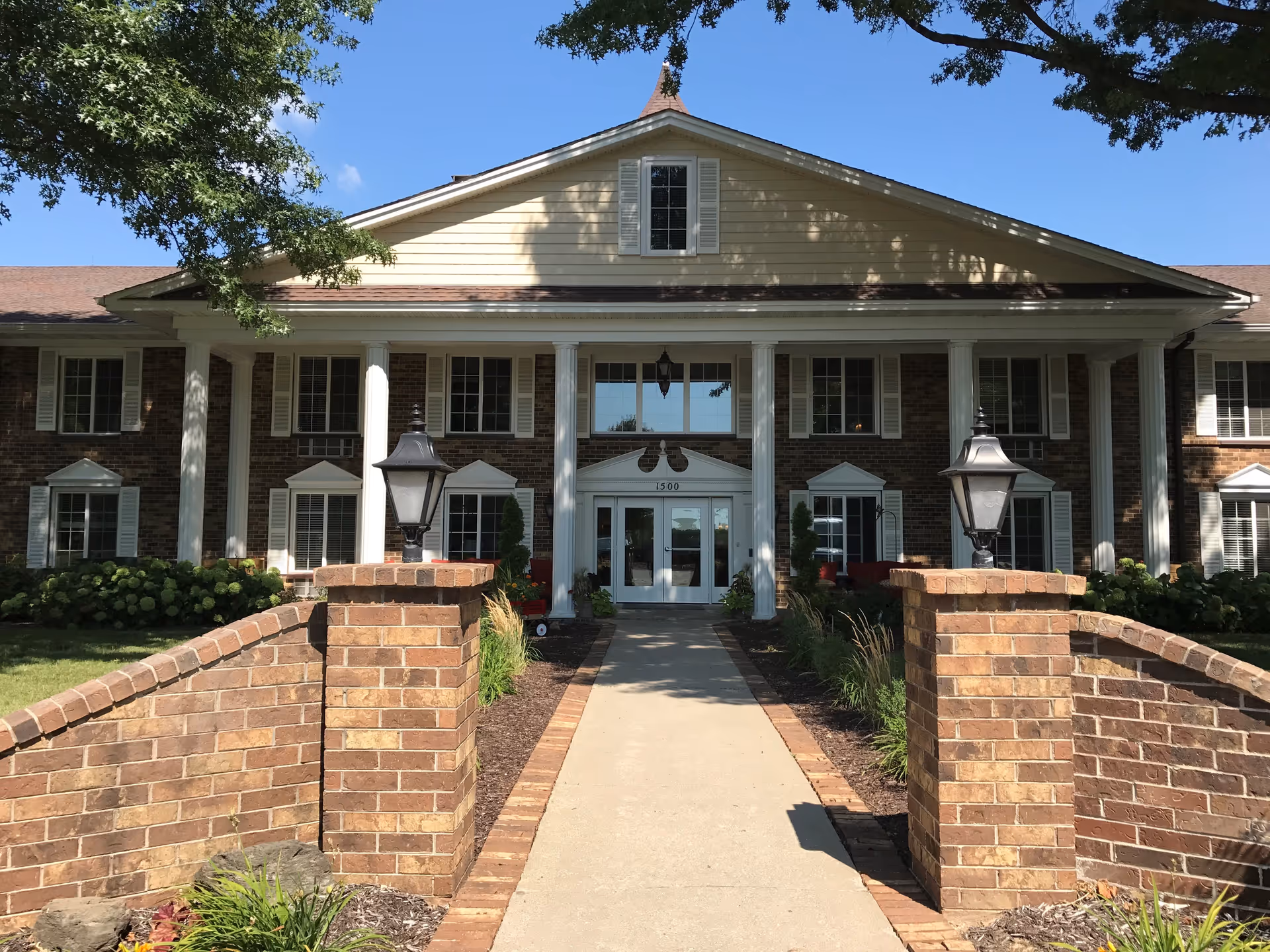 Front exterior view of a two-story brick building with white columns and multiple windows, a concrete walkway leading to double glass doors, flanked by brick pillars with black lanterns, surrounded by greenery and trees under a clear blue sky.