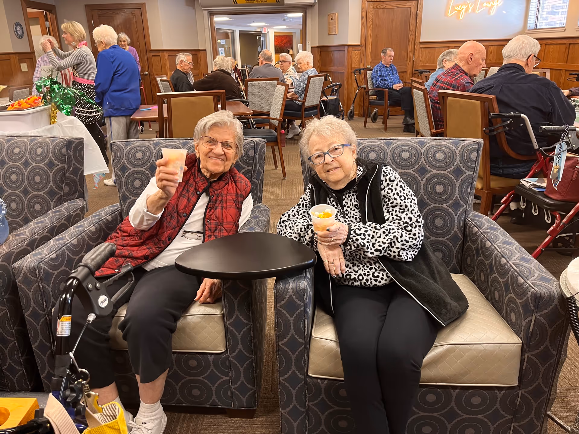 Two elderly women sitting in patterned armchairs in a communal lounge area, each holding a cup with snacks. Behind them, several other elderly people are seated at tables or standing, engaging in conversation. The room has wood paneling on the walls and a warm, inviting atmosphere.