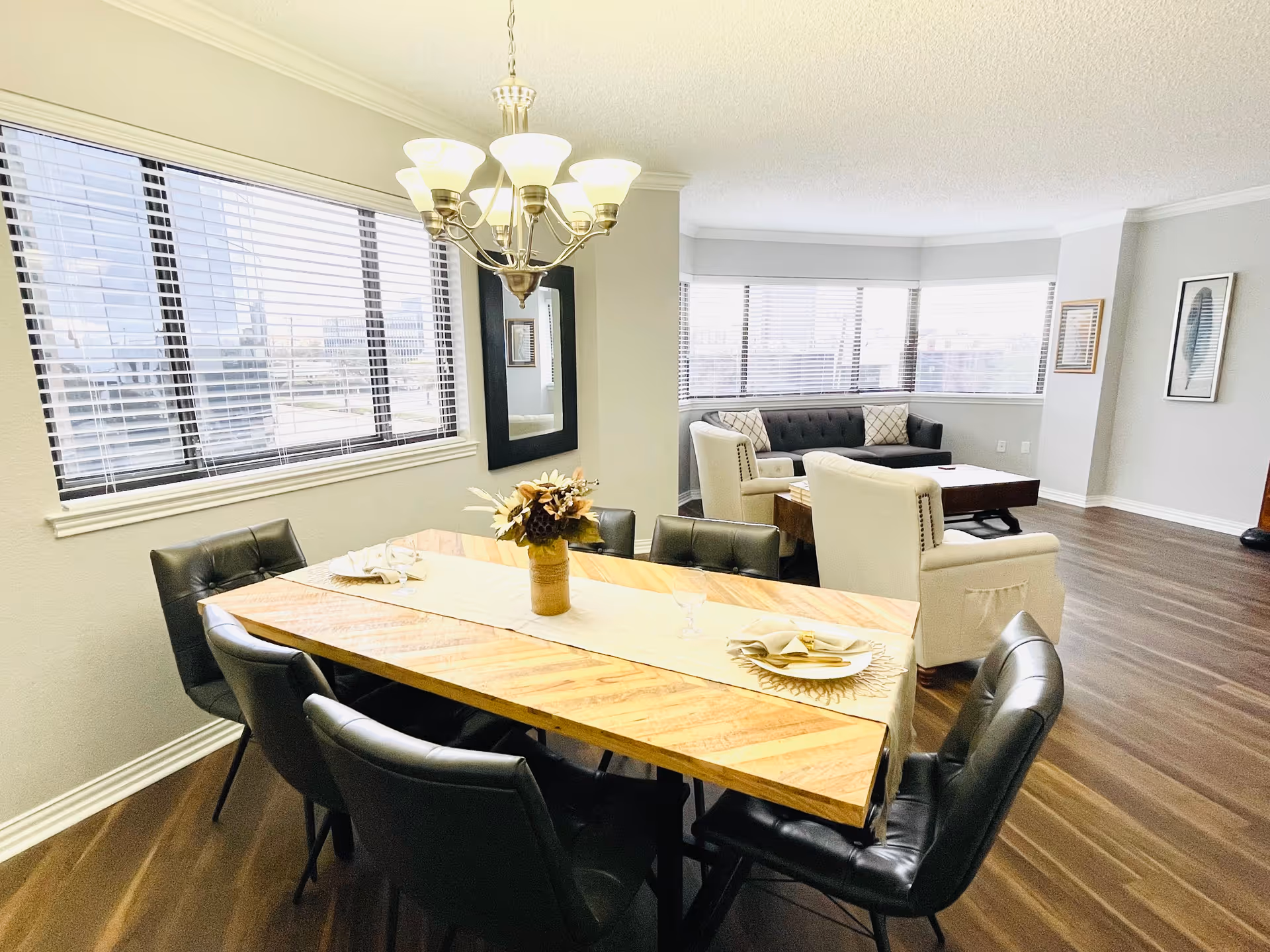 A bright dining and living area with a wooden dining table set for two, surrounded by six black cushioned chairs. A chandelier with multiple lights hangs above the table. In the background, there is a living room with a black sofa, two white armchairs, a wooden coffee table, and large windows with blinds letting in natural light. The floor is wooden, and the walls are painted light gray with framed artwork.