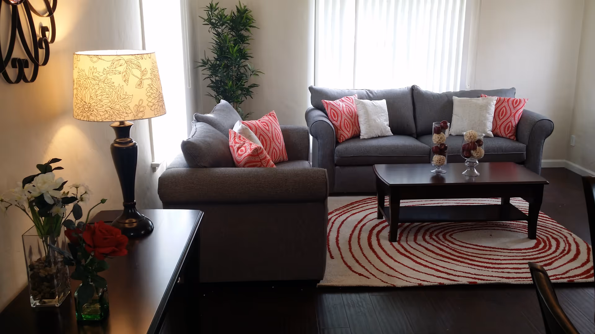 A cozy living room with two gray sofas adorned with red and white patterned pillows. A dark wooden coffee table sits on a white rug with red circular patterns. A side table with a decorative lamp and flower vases is positioned to the left, and a tall green plant is placed near the window with white curtains.