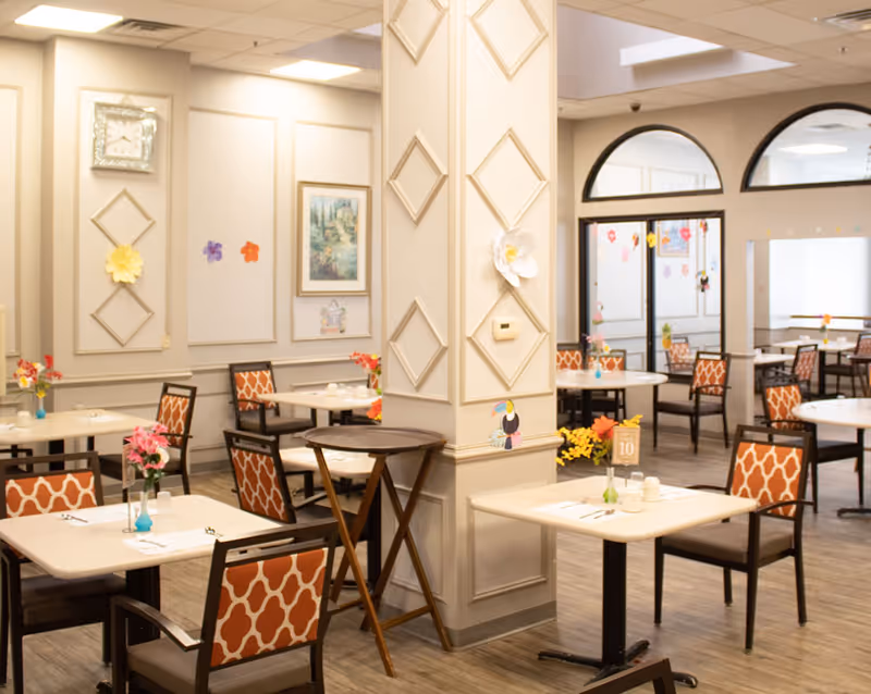 Bright assisted-living dining room with multiple small tables, patterned chairs, floral centerpieces and decorative wall panels.