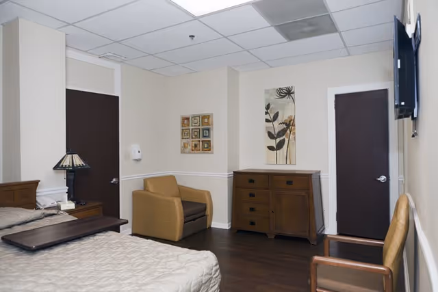 A senior living facility bedroom with a bed covered in a beige quilt, a wooden nightstand with a lamp, a brown armchair, a wooden dresser with drawers, two closed dark brown doors, a wall-mounted TV, and two pieces of wall art featuring floral designs.