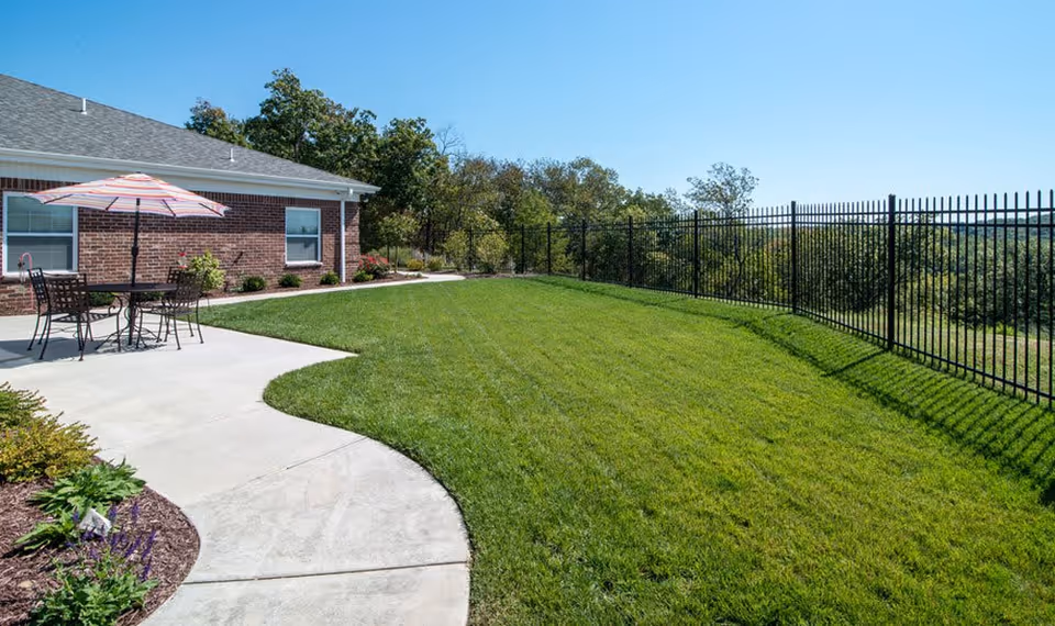 Outdoor patio area at Colony Pointe Senior Living featuring a concrete walkway, a table with four chairs and a striped umbrella, a well-maintained grassy lawn, a black metal fence, and a brick building with windows under a clear blue sky.