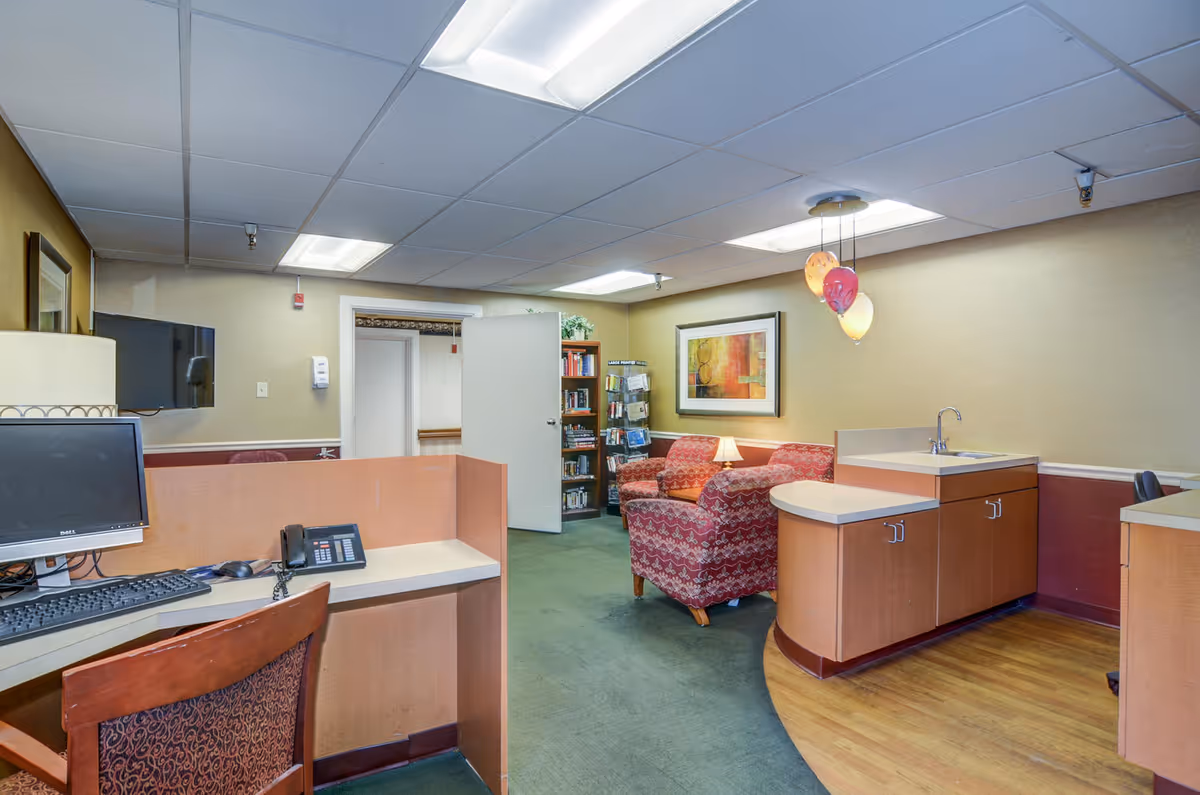Interior view of a healthcare center office area with a computer workstation, a telephone, a red patterned armchair, a small lamp on a side table, a bookshelf filled with books, and a small sink area. The room has beige walls, a green carpet, and a drop ceiling with fluorescent lights. There are three balloons hanging from the ceiling.
