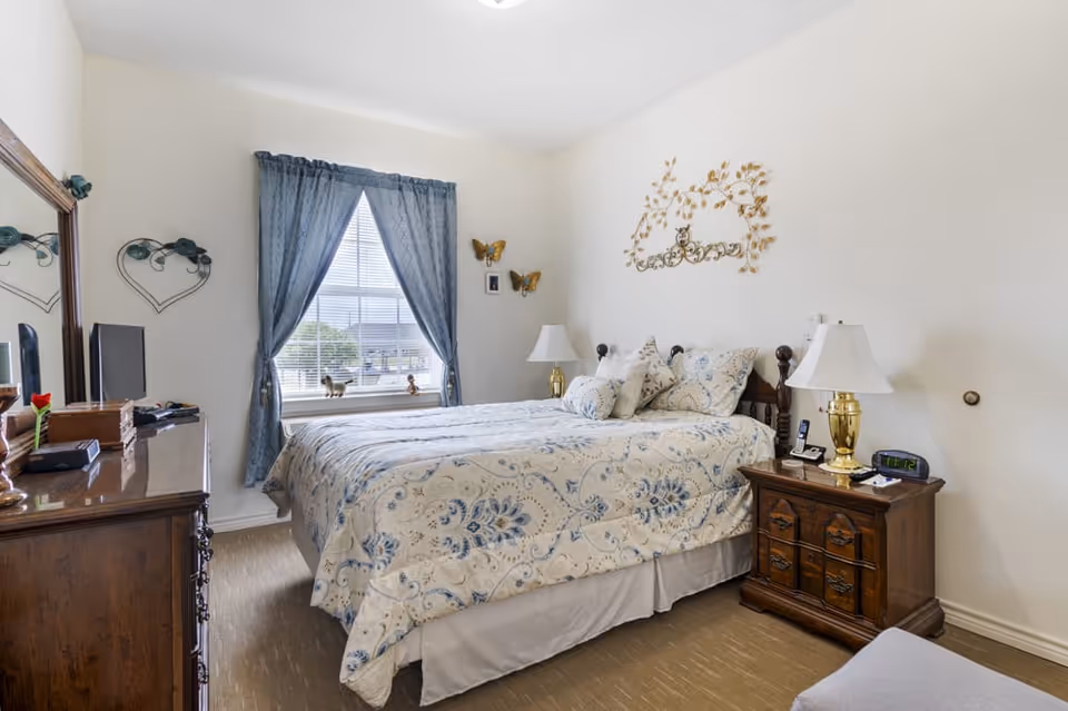 A cozy bedroom with a bed covered in a blue and white patterned comforter and multiple pillows. The room features a wooden nightstand with a lamp, alarm clock, and phone, and a wooden dresser with a mirror and TV on top. Blue curtains frame a window letting in natural light, and decorative wall art including butterflies and a floral metal piece adorn the walls.
