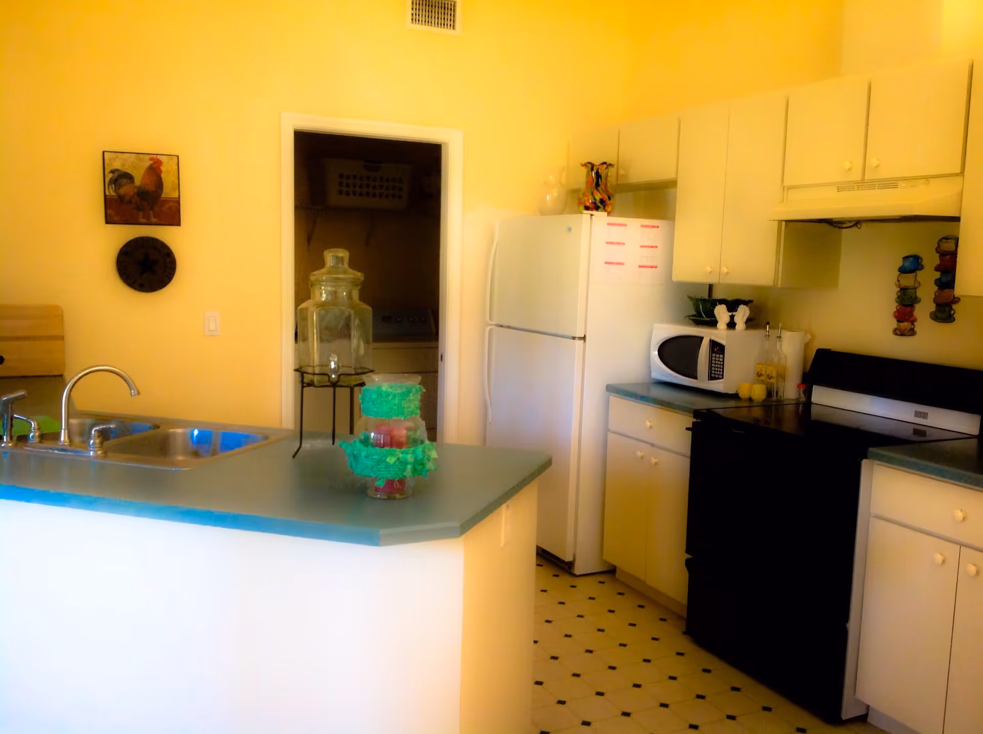 A kitchen area with white cabinets, a black stove, a white refrigerator, and a microwave on the countertop. There is a double sink on the kitchen island with a glass beverage dispenser and a decorative item on the counter. The walls are painted yellow, and there is a doorway leading to a laundry room with a washing machine and a laundry basket visible. The floor has a white and black patterned linoleum.